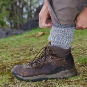 A person's foot in a brown boot resting on green grass, with a gray sock extending out of the top of the boot. The person is tucking his brown pants leg into the sock.